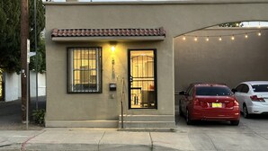 A two-story building with a beige exterior and a red-tiled roof. There is a black-framed window with a metal grill, a door with a glass panel, and a small light fixture above the door. Two cars are parked in the driveway. - Cozy Retro Downtown Living (Albuquerque)