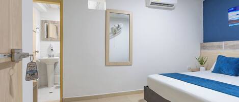 A hotel room with a bed, a mirror, an air conditioning unit, a sink, and a door leading to a bathroom.