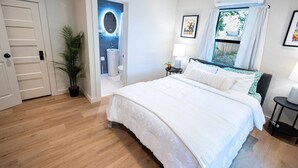 Habitación de hotel con una cama grande, baño con espejo, planta en maceta y ventana con cortinas.