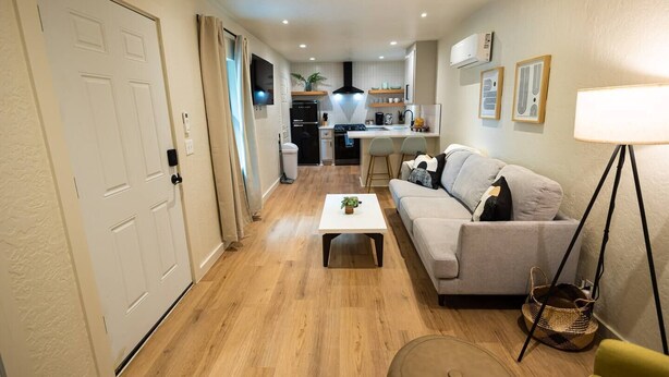 Un espacio habitable compacto con un sofá, una mesita de centro y una lámpara. Se ve un área de cocina al fondo con un refrigerador y un lavamanos.