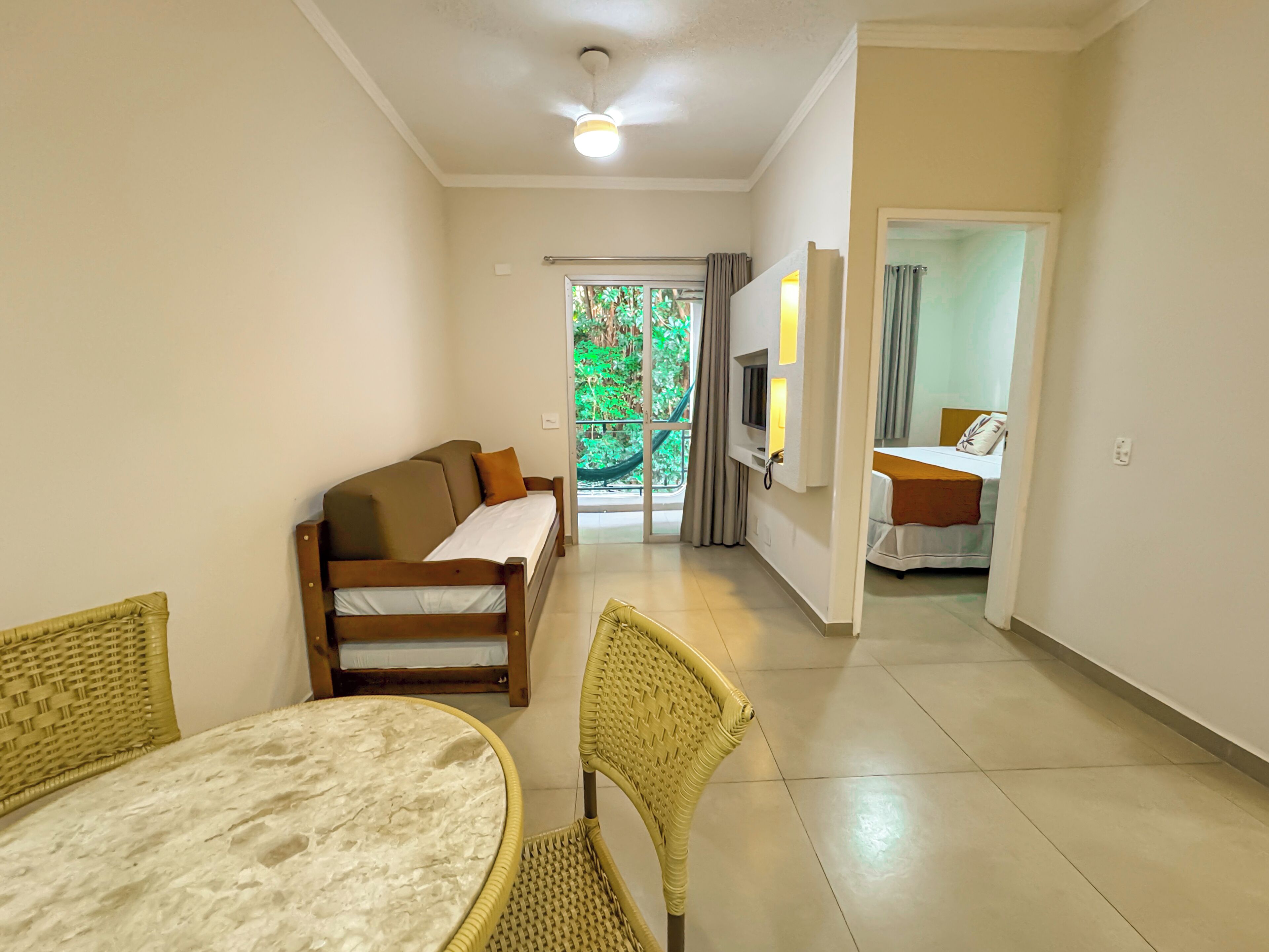 Quarto de hotel com sofá, mesa de jantar, cadeiras de vime, televisão e varanda com vista para o verde.