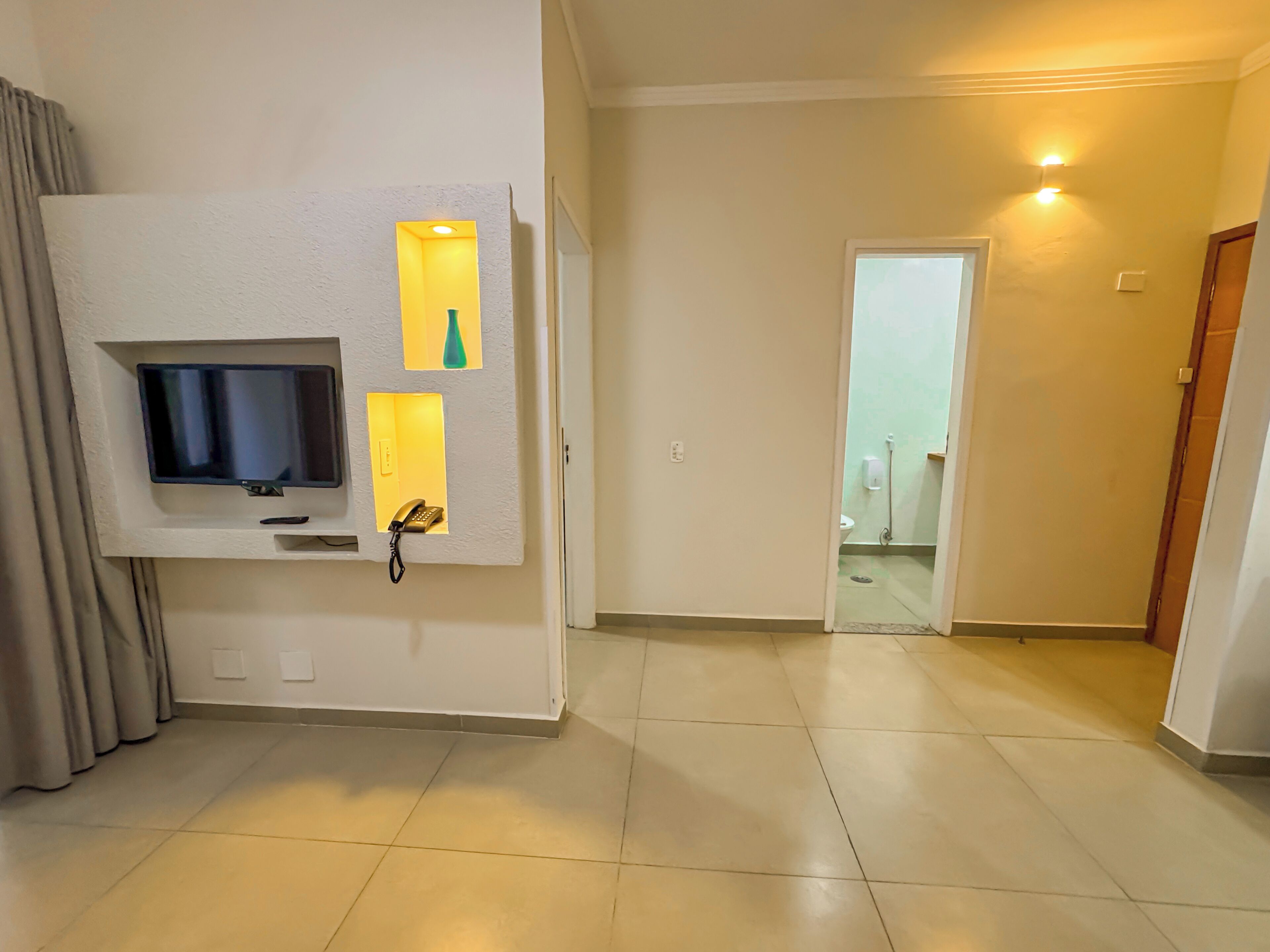 A hotel room with a flat-screen TV mounted on the wall, a telephone, and a small shelf with a vase. The room has a tiled floor and a door leading to a bathroom.