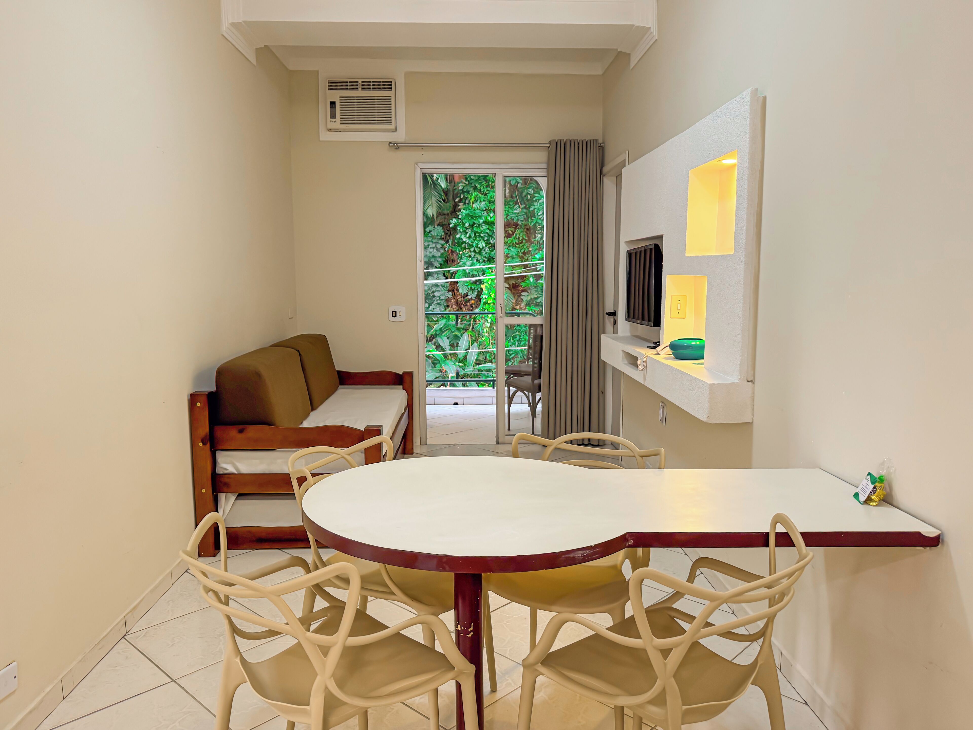 A compact living space with a round dining table, beige chairs, a sofa, a TV, and a small kitchen area.