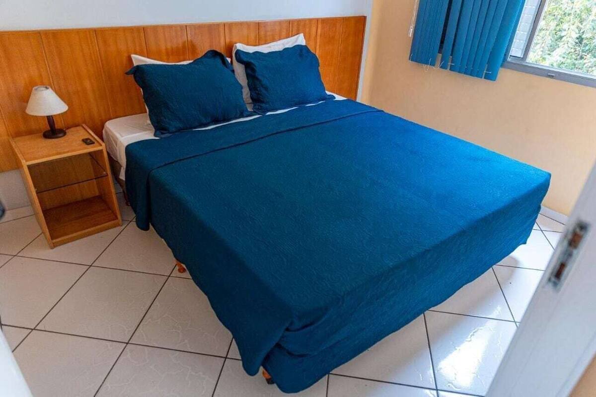 Ein Bett mit blauer Bettdecke, einem Holzkopfschild, einem Nachttisch mit Lampe und einem Fenster mit Vorhängen.