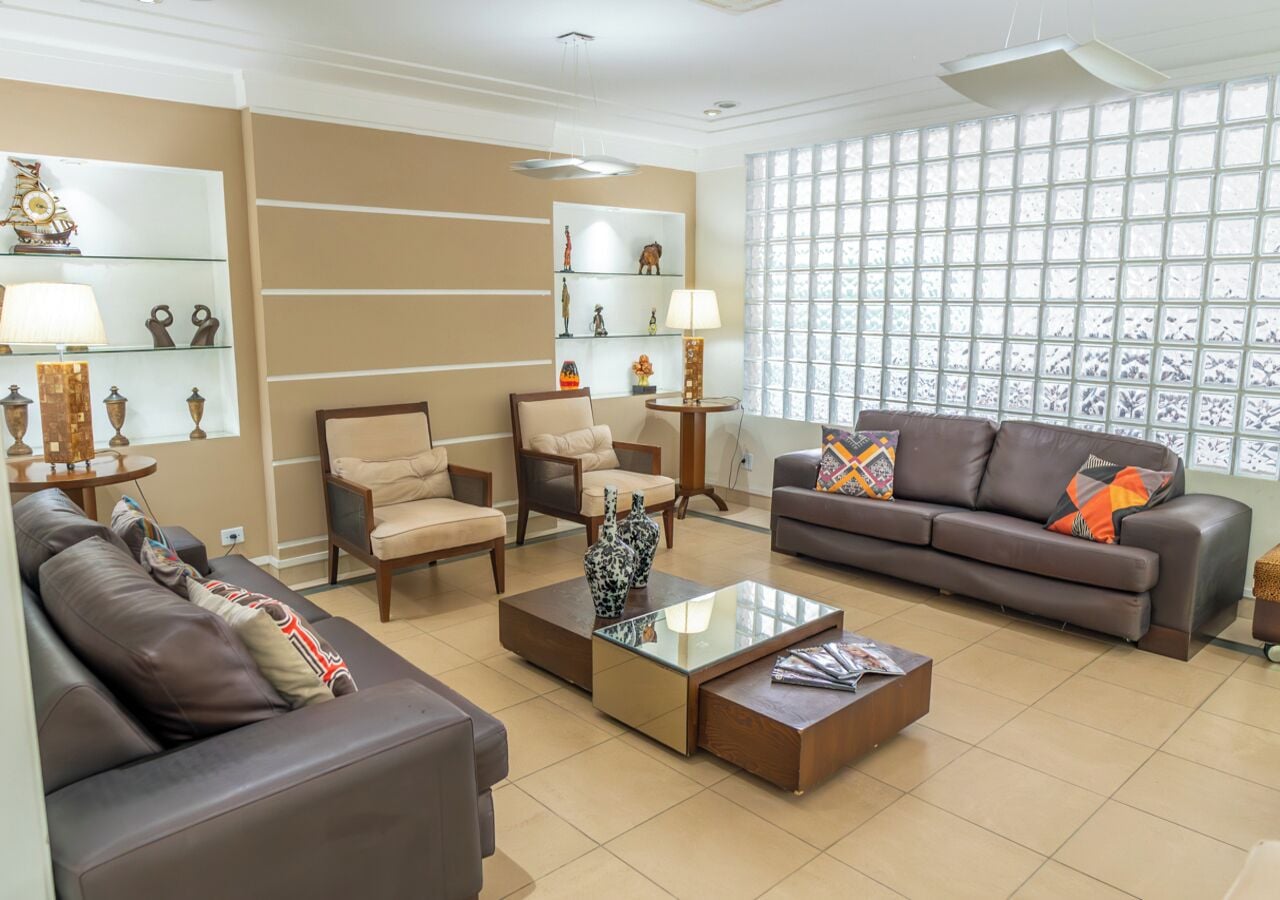 A spacious living room with a brown leather sofa, beige armchairs, and a brown leather couch. There is a glass coffee table with decorative vases. The room has a large window with a view of the outside.