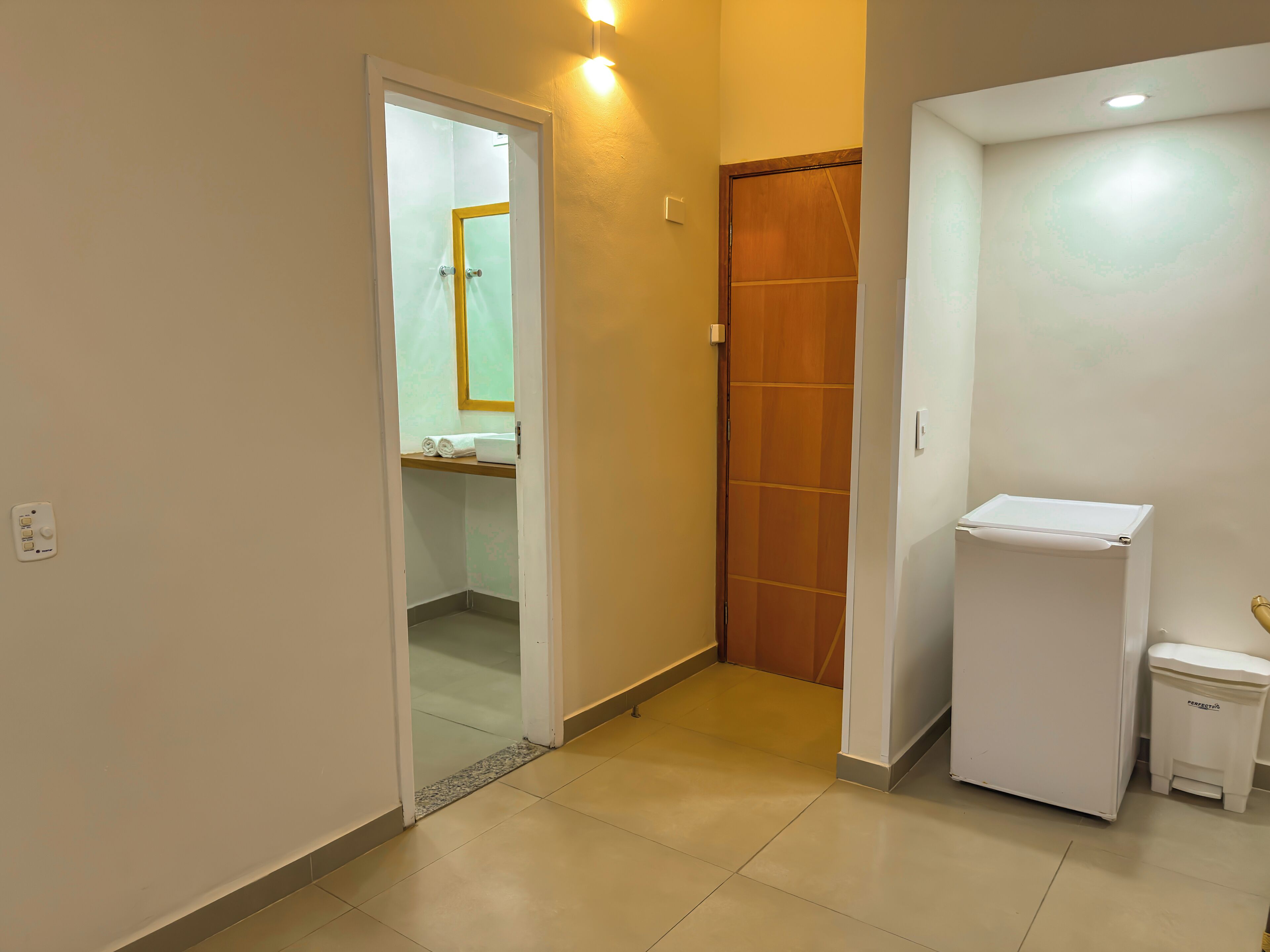 A hallway with a door leading to a bathroom, a small refrigerator, and a trash bin.