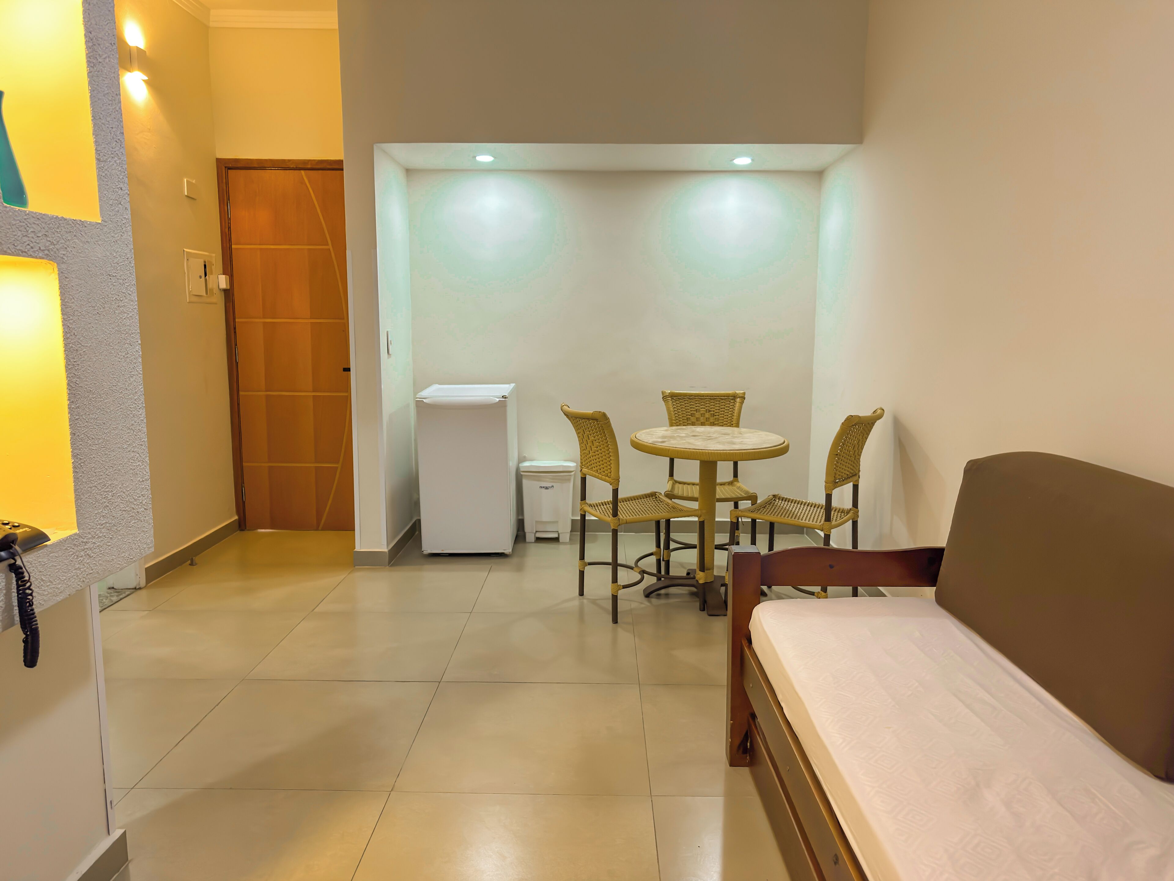 Habitación de hotel con una cama, una mesita pequeña con sillas, un cesto de basura y una puerta.