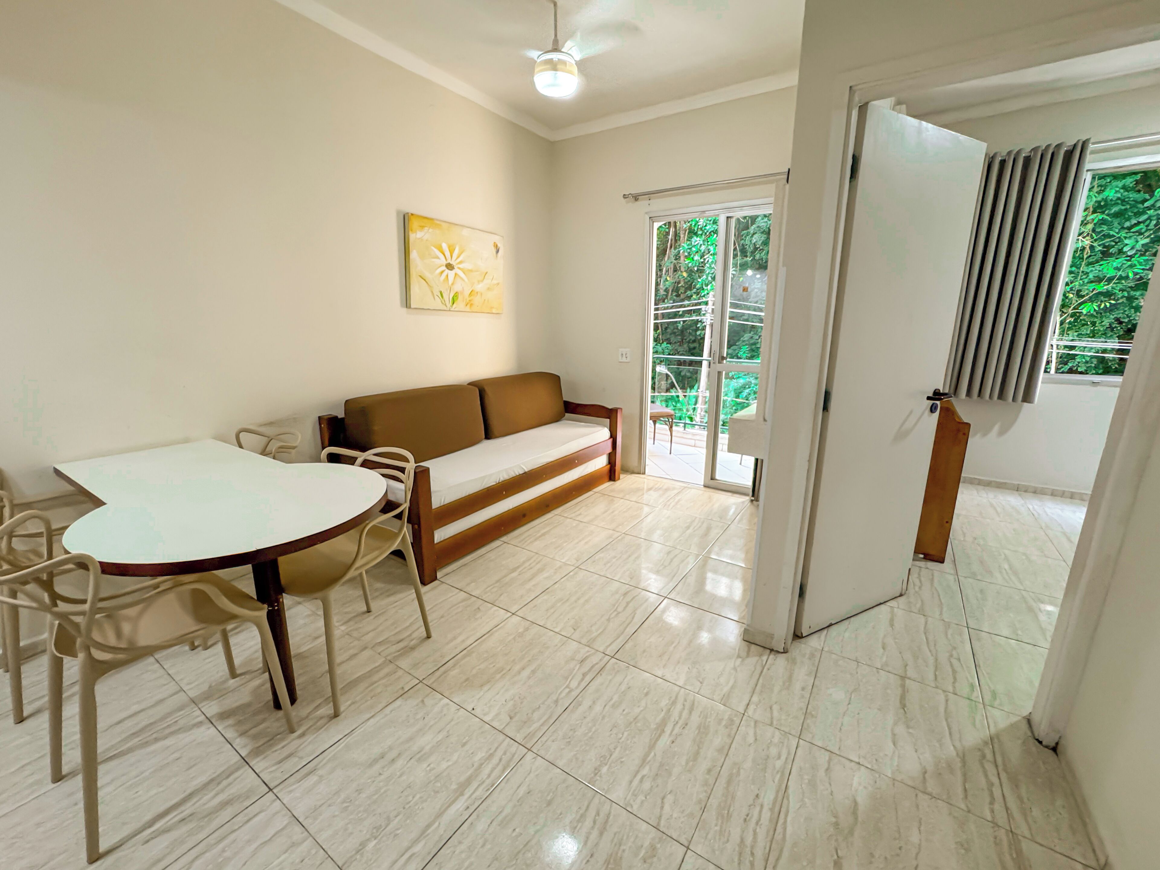 A living room with a sofa, a dining table, and chairs. There is a sliding glass door leading to a balcony with a view of greenery.