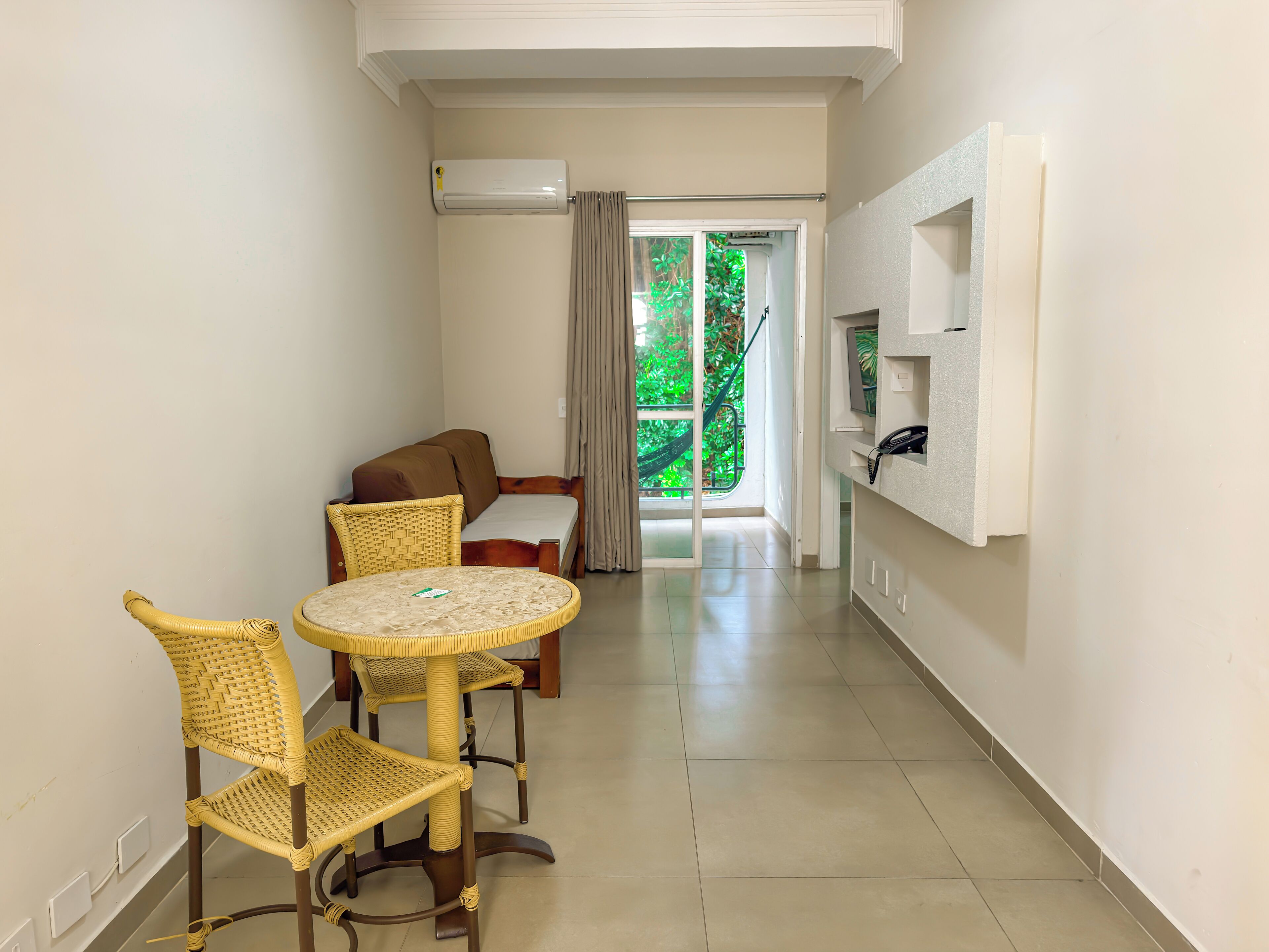 Une pièce avec une table ronde et deux chaises en osier, un canapé, un téléviseur fixé au mur et une vue sur la verdure à l’extérieur.