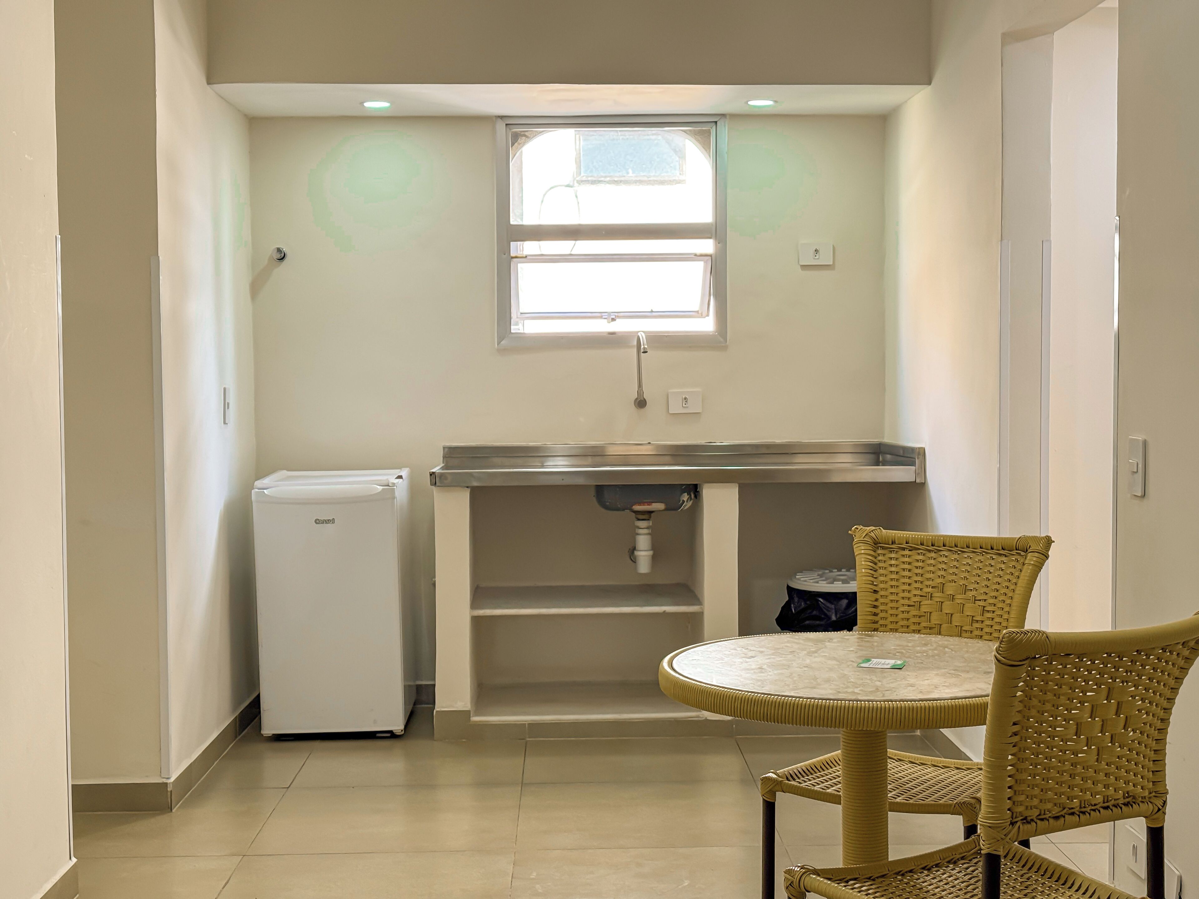 A compact kitchen with a refrigerator, a sink, and a window above it. There is a round table with a chair in front of the sink.