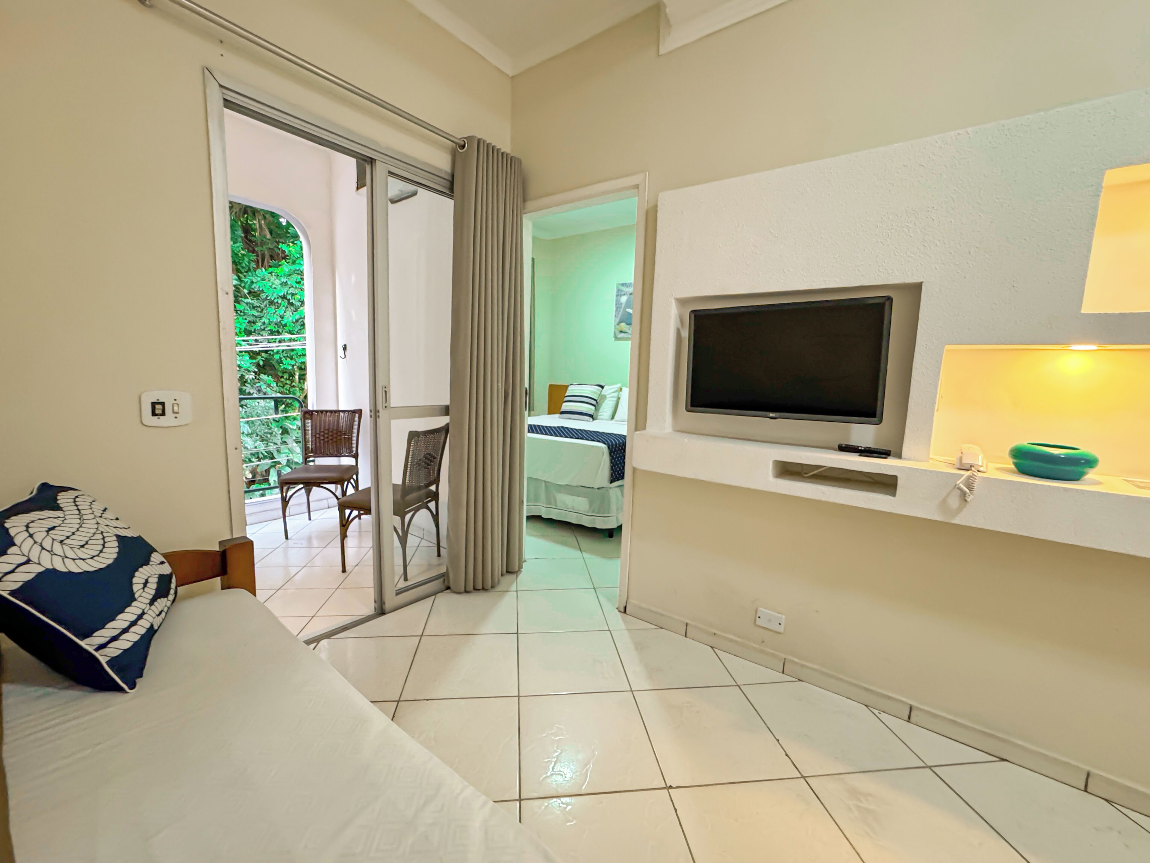 A hotel room with a bed, a TV, a lamp, and a view of a balcony with chairs.