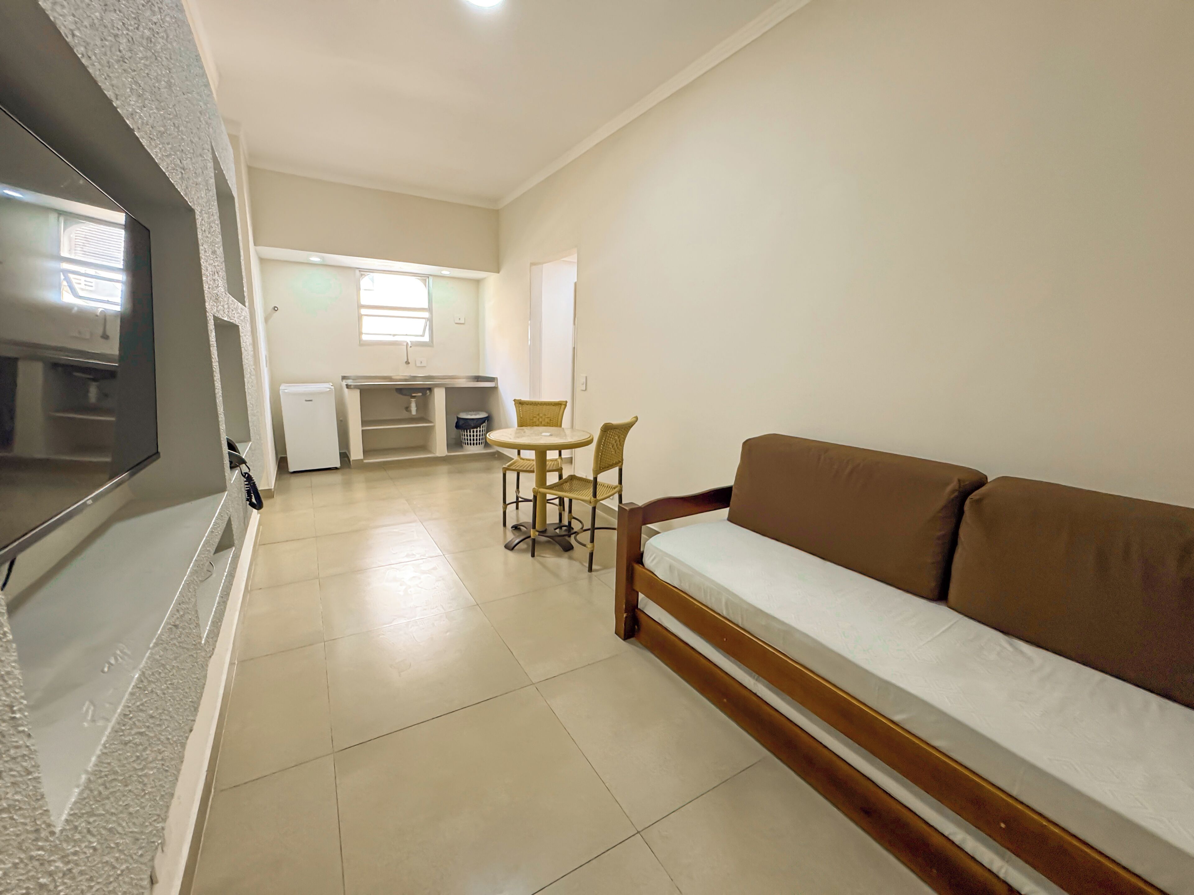 A living room with a sofa, a dining table with chairs, a kitchen area, and a TV mounted on the wall.