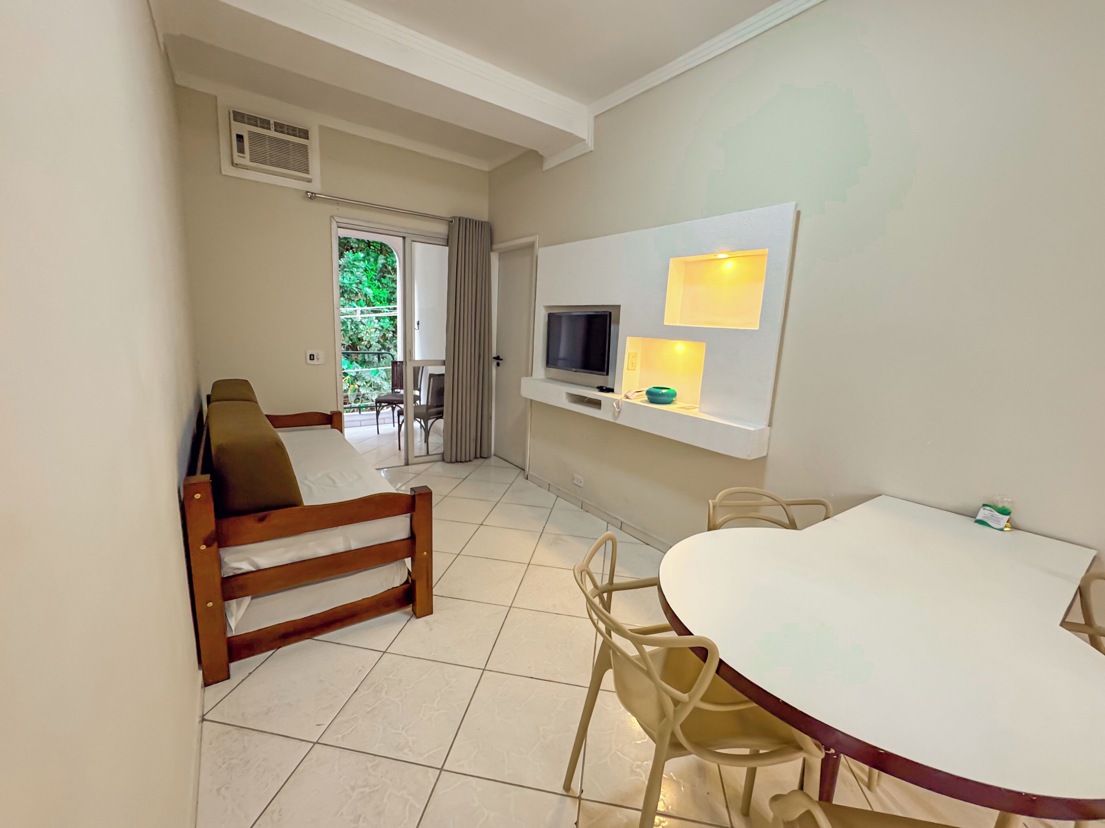 Una habitación con una cama, una mesa de comedor con sillas, un televisor montado en la pared y un ventanal con cortinas.
