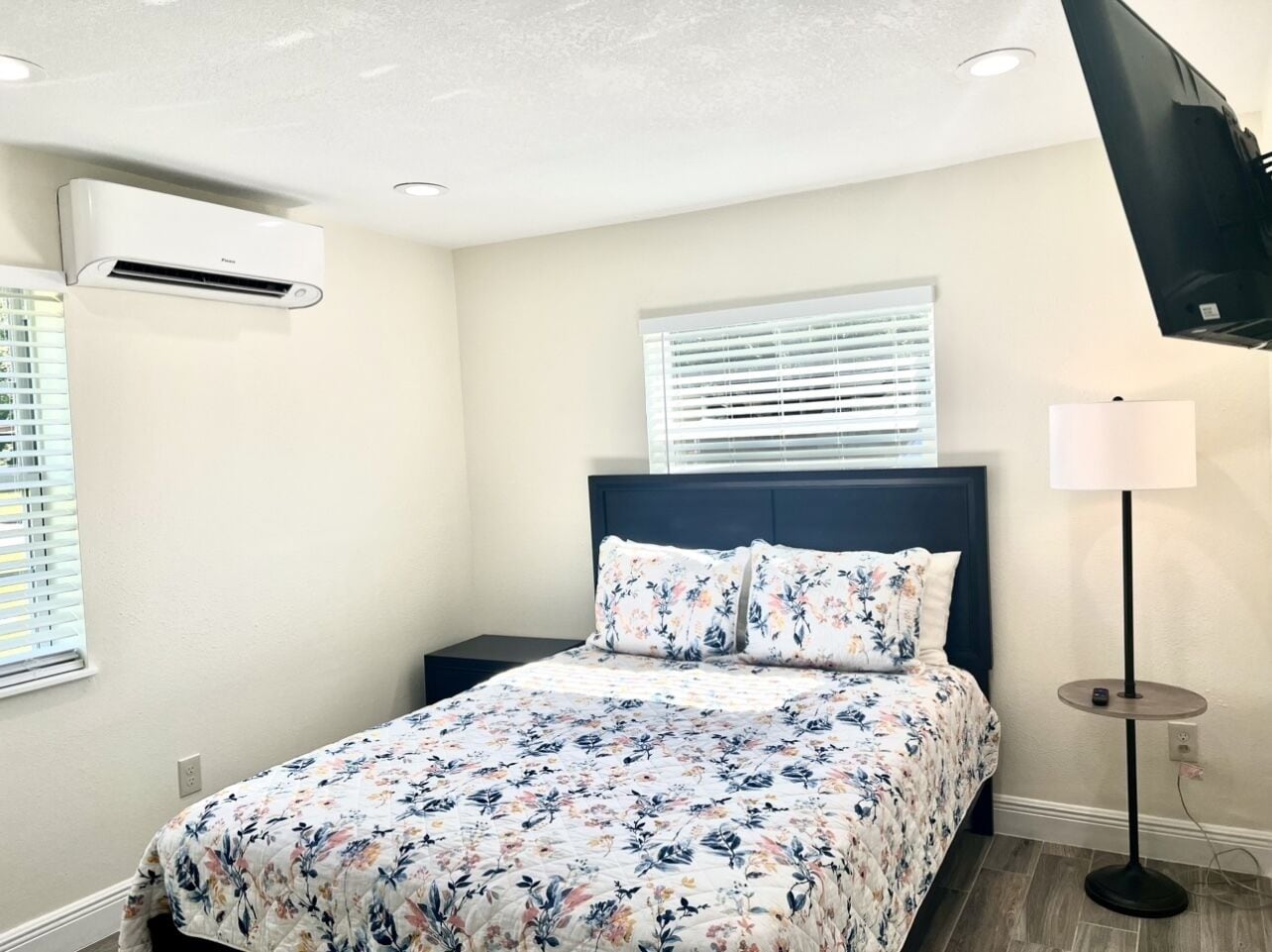 Un dormitorio con cama, mesita de noche, lámpara, ventana con persianas y unidad de aire acondicionado.