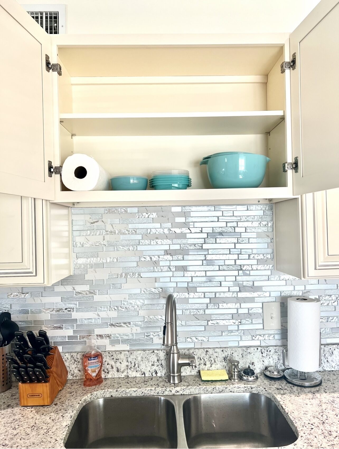 A kitchen with a double sink, a stainless steel faucet, and a backsplash made of white and gray tiles. There are cabinets with open doors displaying blue bowls and a roll of paper towels.