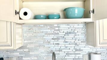 A kitchen with a double sink, a stainless steel faucet, and a backsplash made of white and gray tiles. There are cabinets with open doors displaying blue bowls and a roll of paper towels.