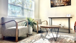 A living room with two patterned armchairs, a wooden coffee table, a potted plant, a large window with blinds, a wall-mounted TV, and a small shelf with decorative items. - Home Sweet Home (Sanford)