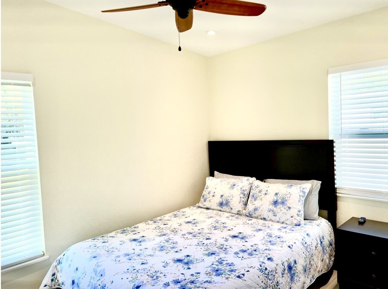 Une chambre à coucher avec un lit, un ventilateur de plafond et deux fenêtres équipées de stores.