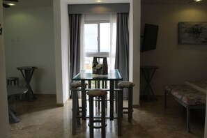 Un coin repas comprenant une table à plateau en verre et quatre chaises, un banc, et une vue par une fenêtre agrémentée de rideaux.