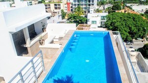Una piscina en la azotea con agua cristalina azul, rodeada por un edificio blanco y vistas de la ciudad.