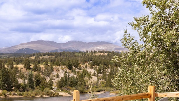 Two Adirondack chairs facing a river with a wooden fence and a mountainous backdrop. - The Hand Hotel (Fairplay)