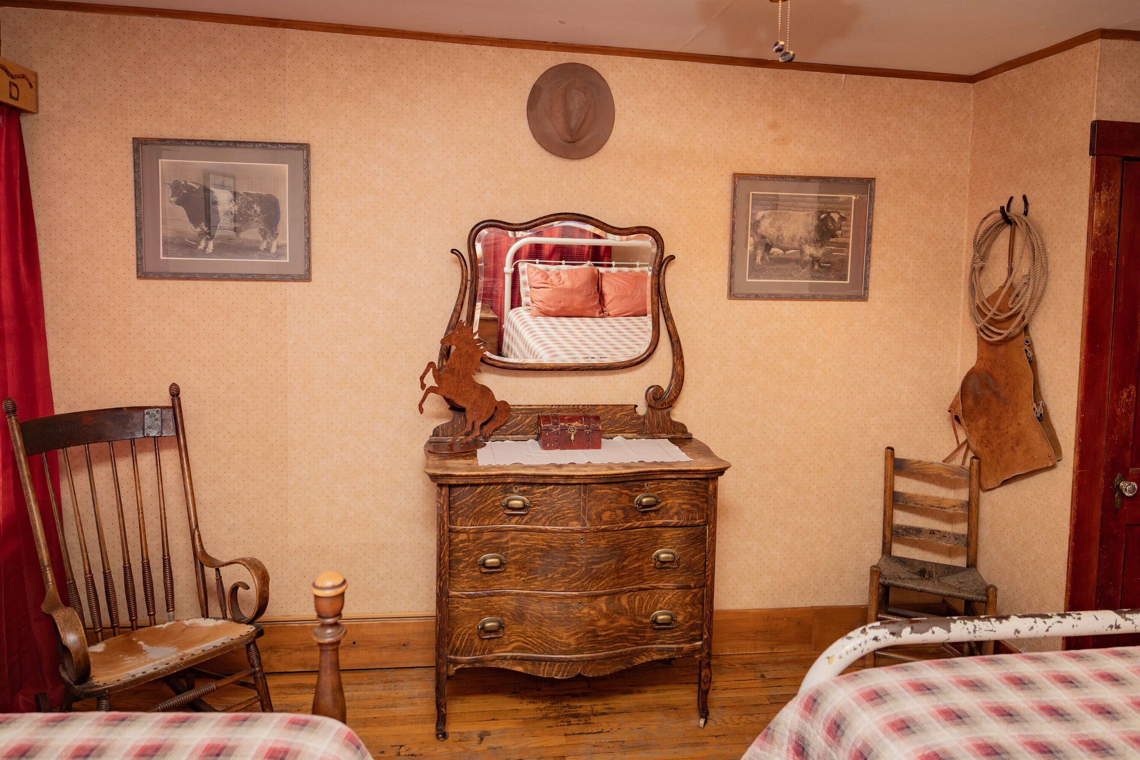A room with a wooden dresser, a bed with a checkered bedspread, a rocking chair, and two framed pictures on the wall.