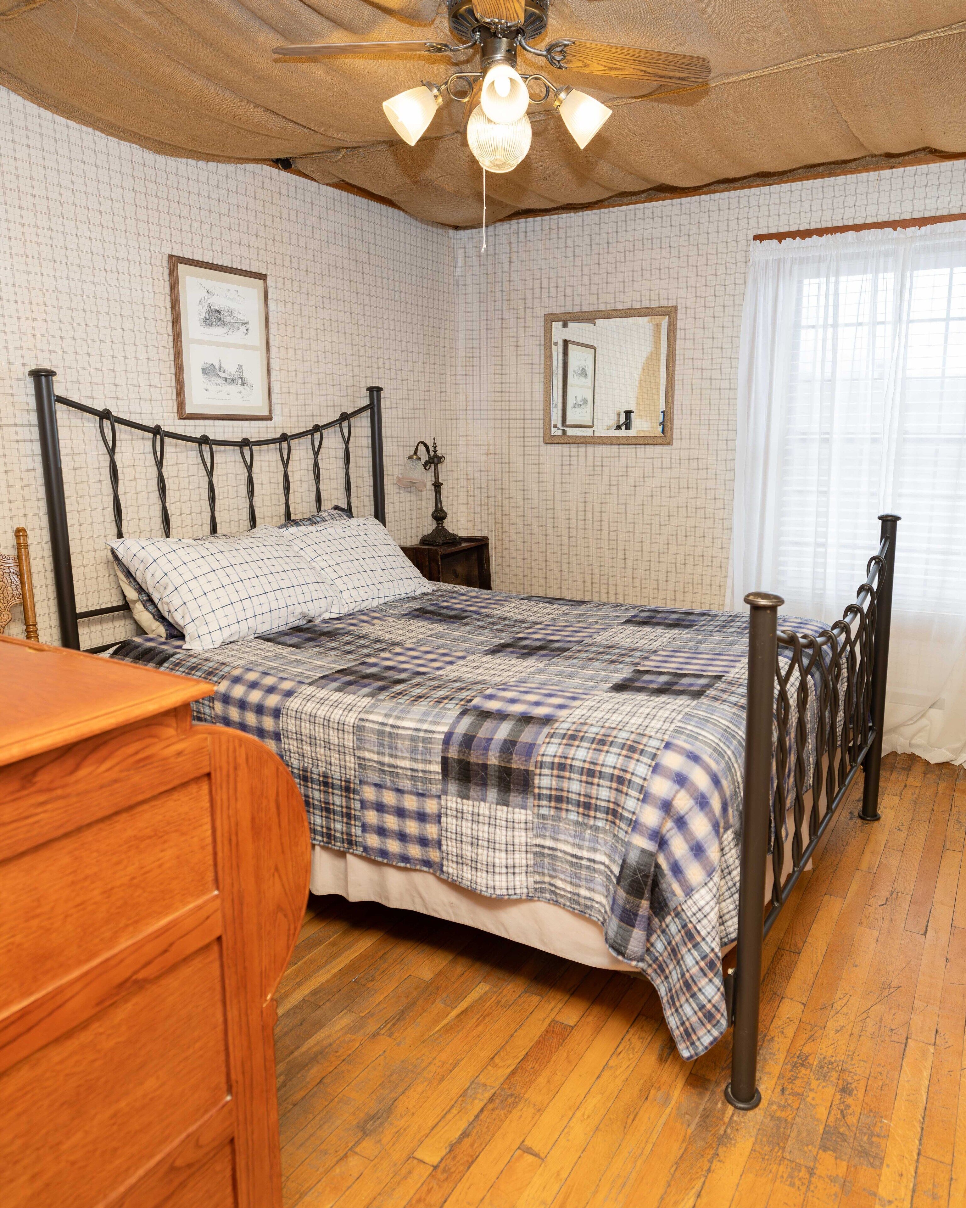 Un dormitorio con cama, piso de madera, ventilador de techo y ventana con cortinas.