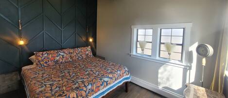 Quarto com cama, janela com cortinas, mesa de cabeceira e luminária na parede.
