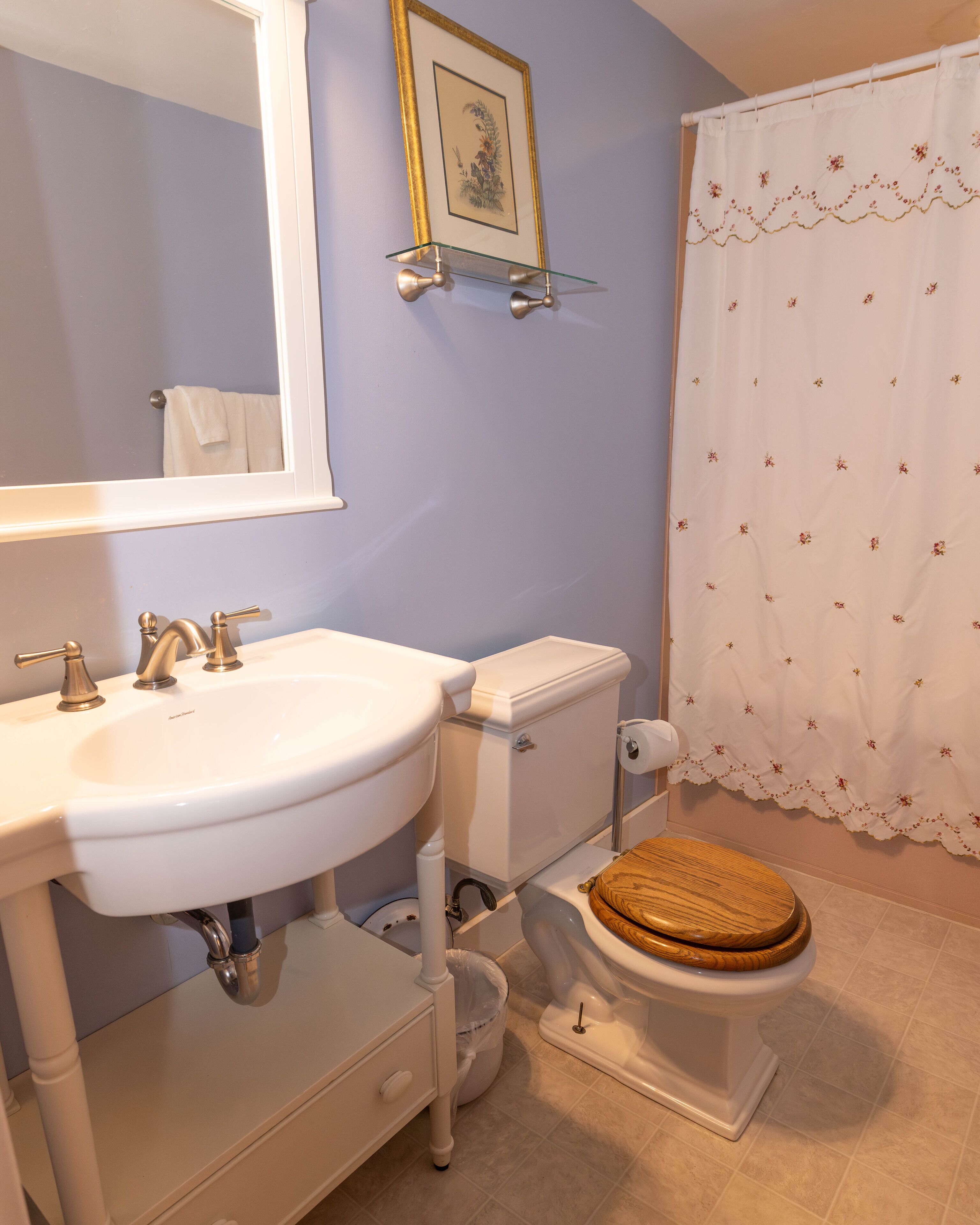 Ein Badezimmer mit einem weißen Waschbecken, einer Toilette mit Holzsitzen, einem gerahmten Bild an der Wand und einem Duschvorhang mit Blumenmuster.
