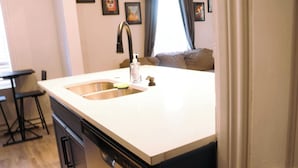 A kitchen with a white countertop, stainless steel sink, and a dishwasher. There is a bar stool and a window with curtains. - Near Riverwalk, Hemisfair, Art & Dining Spots (San Antonio)