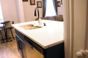 A kitchen with a white countertop, stainless steel sink, and a dishwasher. There is a bar stool and a window with curtains.