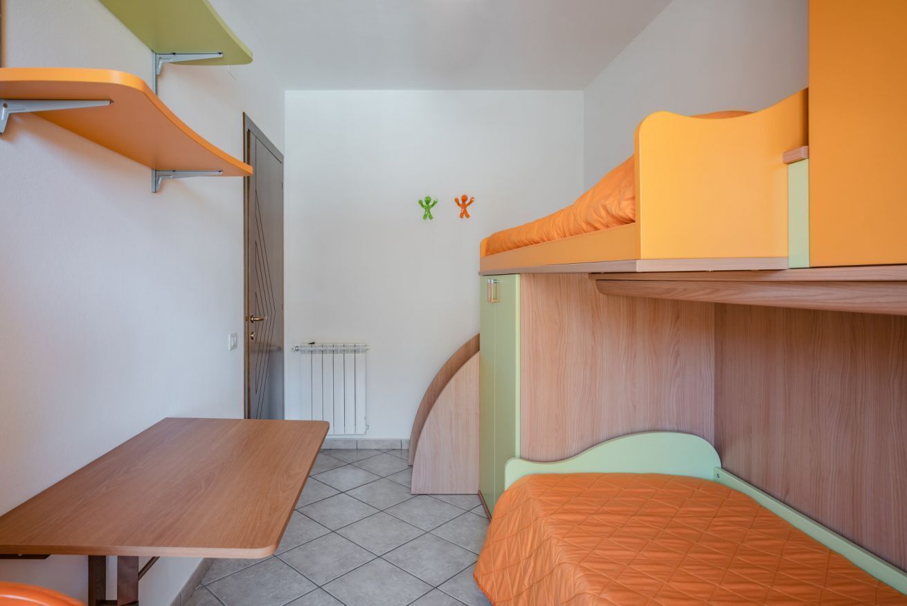 Een kleine, helder verlichte kamer met een stapelbed, een enkel bed, een bureau en een radiator.