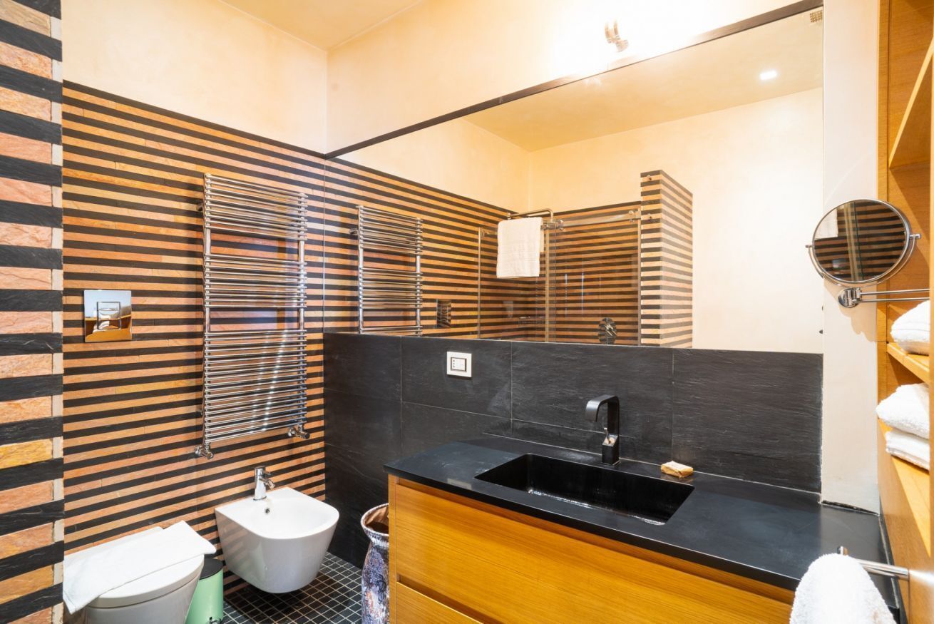A modern bathroom with a black sink, white pedestal sink, and a bidet. The walls have a striped pattern, and there is a mirror on the wall.