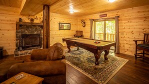 A cozy cabin interior with a fireplace, a pool table, a sofa, and a wooden ceiling.