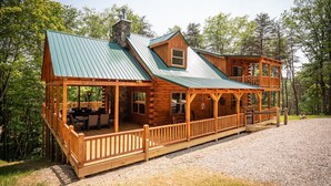 A log cabin with a green metal roof, a stone chimney, and a wooden deck with outdoor seating.