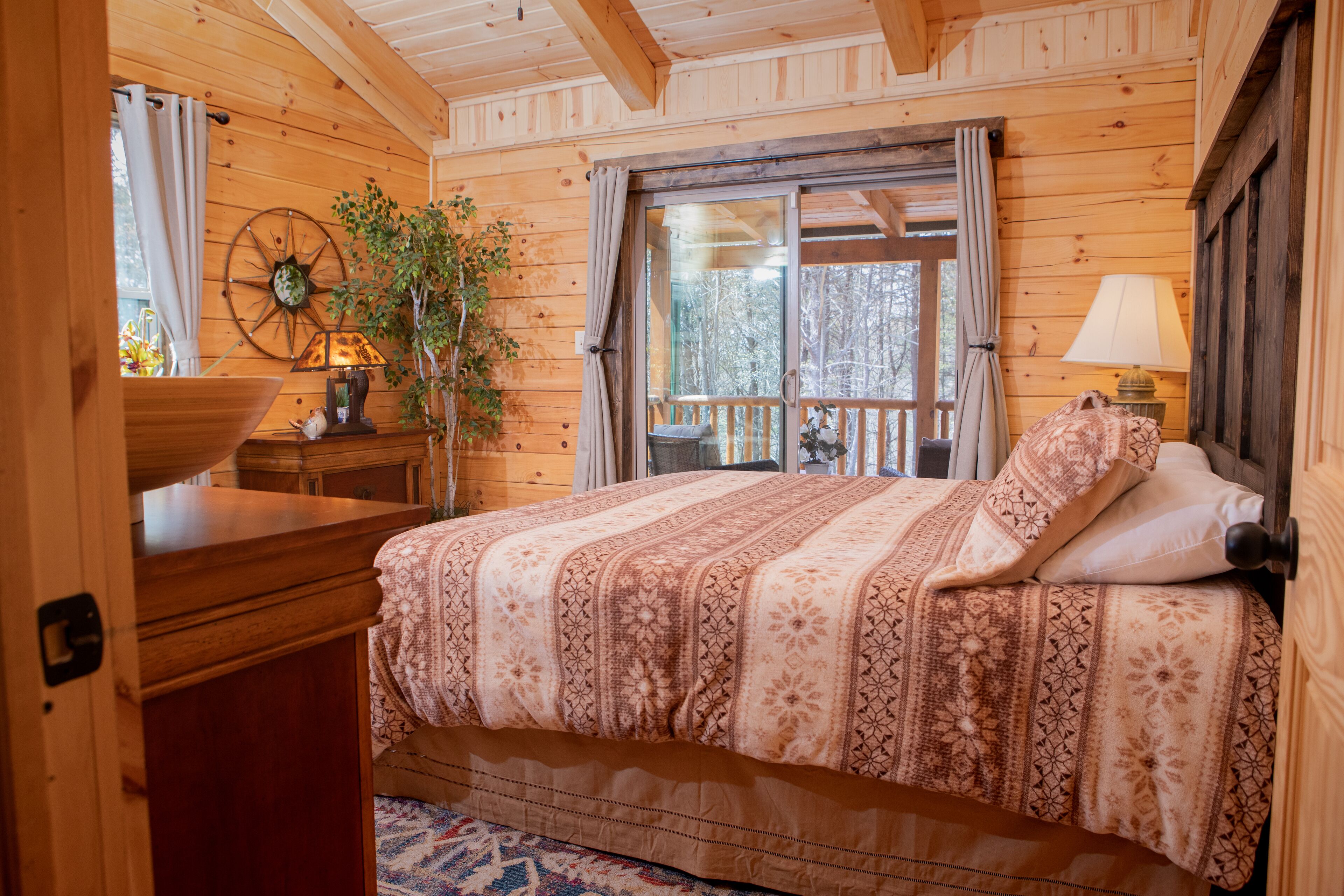 A wooden cabin bedroom with a bed, a nightstand, a lamp, a window with curtains, and a wooden ceiling.
