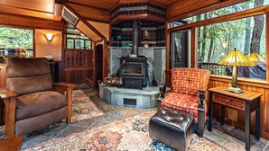 Ein gemütliches Zimmer mit Kamin aus Stein, Holzmöbeln und großen Fenstern mit Blick auf einen Wald.