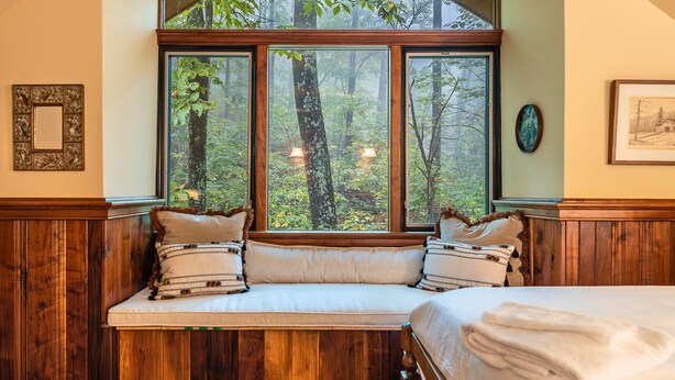 Ein Schlafzimmer mit einem großen Fenster und Blick auf einen Wald. Ein Bett mit weißer Bettwäsche und eine Bank mit Kissen sind vorhanden.