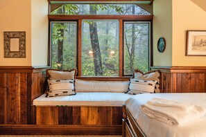 Une chambre avec une grande fenêtre donnant sur une forêt. On y trouve un lit avec une literie blanche et un banc agrémenté de coussins.
