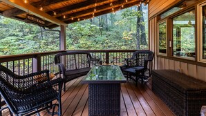 Eine Holzterrasse mit Korbmöbeln und einem Tisch mit Glasplatte, mit Blick auf ein bewaldetes Gebiet.
