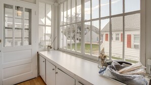 Une cuisine avec des meubles blancs, une grande fenêtre à carreaux et une vue sur les maisons voisines.