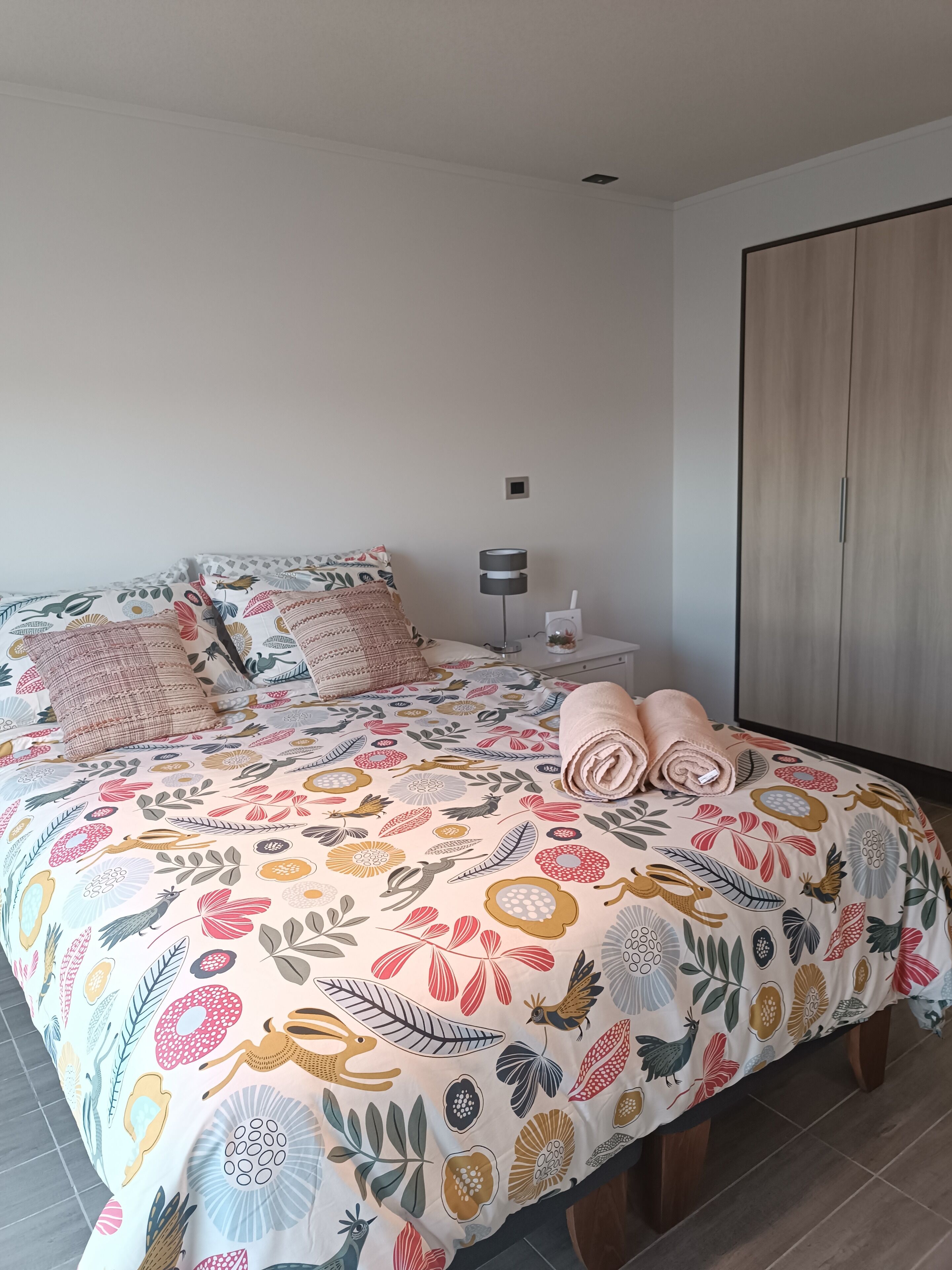 Un dormitorio con edredón estampado con motivos florales, una lámpara de noche y un armario de madera.