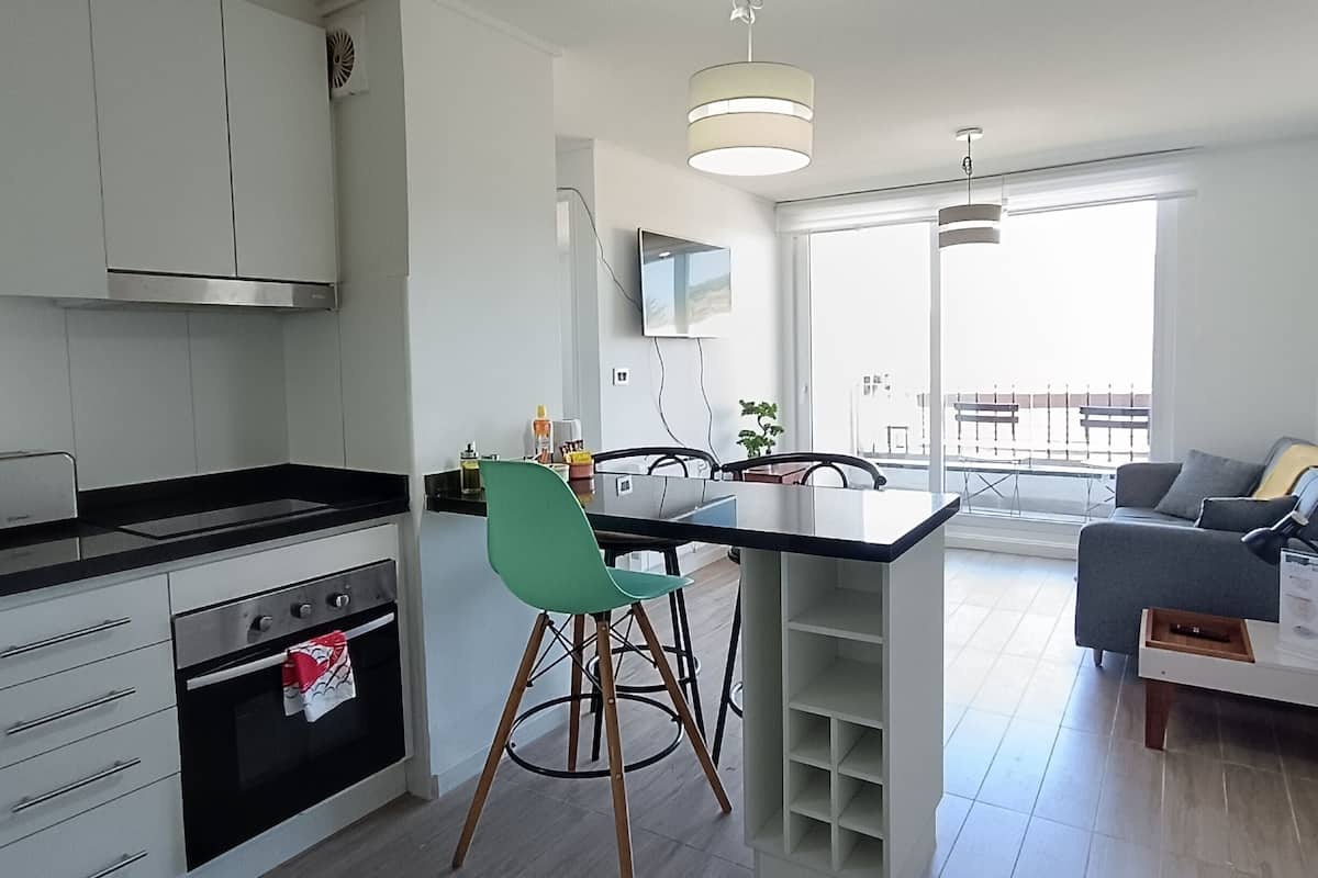 Een moderne keuken met witte kastjes, een zwart aanrecht en een groene stoel. Er is een eethoek met een tafel en stoelen, en een grijze bank in de woonkamer.