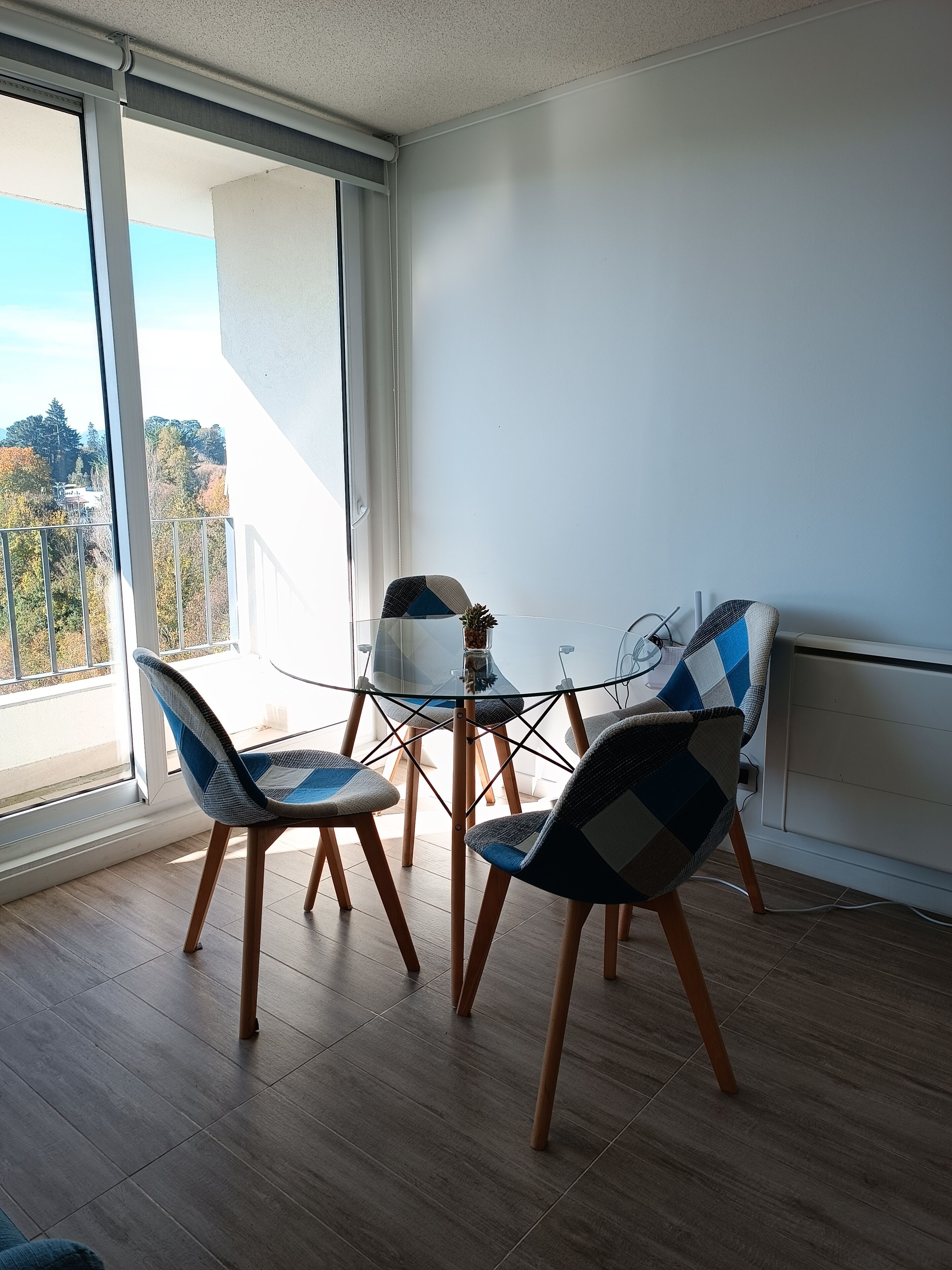 A balcony with a glass table and chairs, wooden flooring, and a view of trees and buildings.