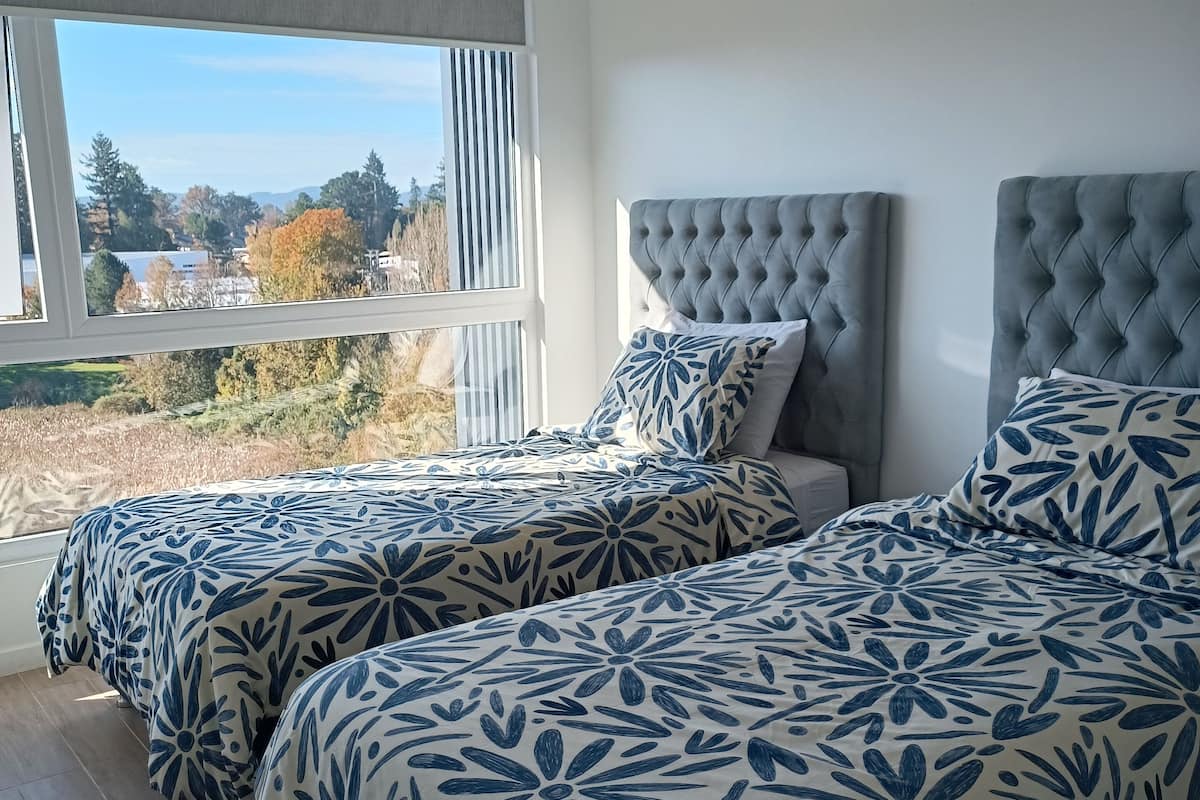 Een slaapkamer met twee bedden, een groot raam en uitzicht op een park en gebouwen.