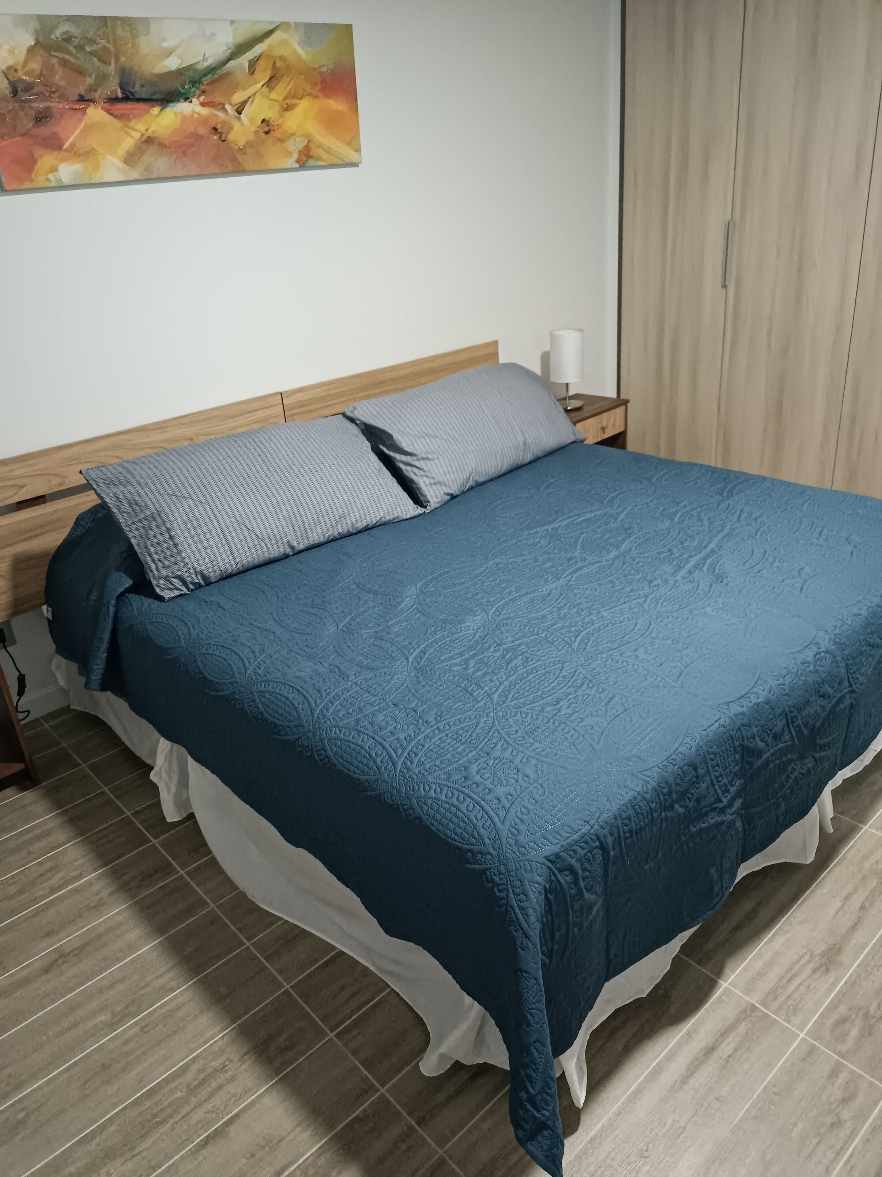 A neatly made bed with a blue bedspread and two pillows, a wooden headboard, a bedside table with a lamp, and a painting on the wall.