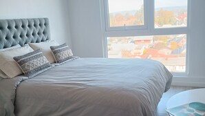 A bedroom with a large bed, a view of a cityscape through a window, and a small round table with a colorful pattern.