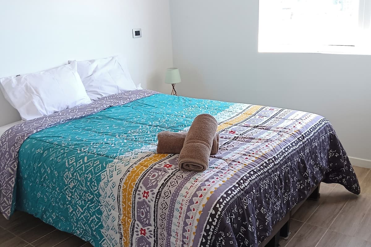 Een netjes opgemaakt bed met beddengoed met een patroon en een opgerolde handdoek, een bedlampje en uitzicht buiten door een raam.