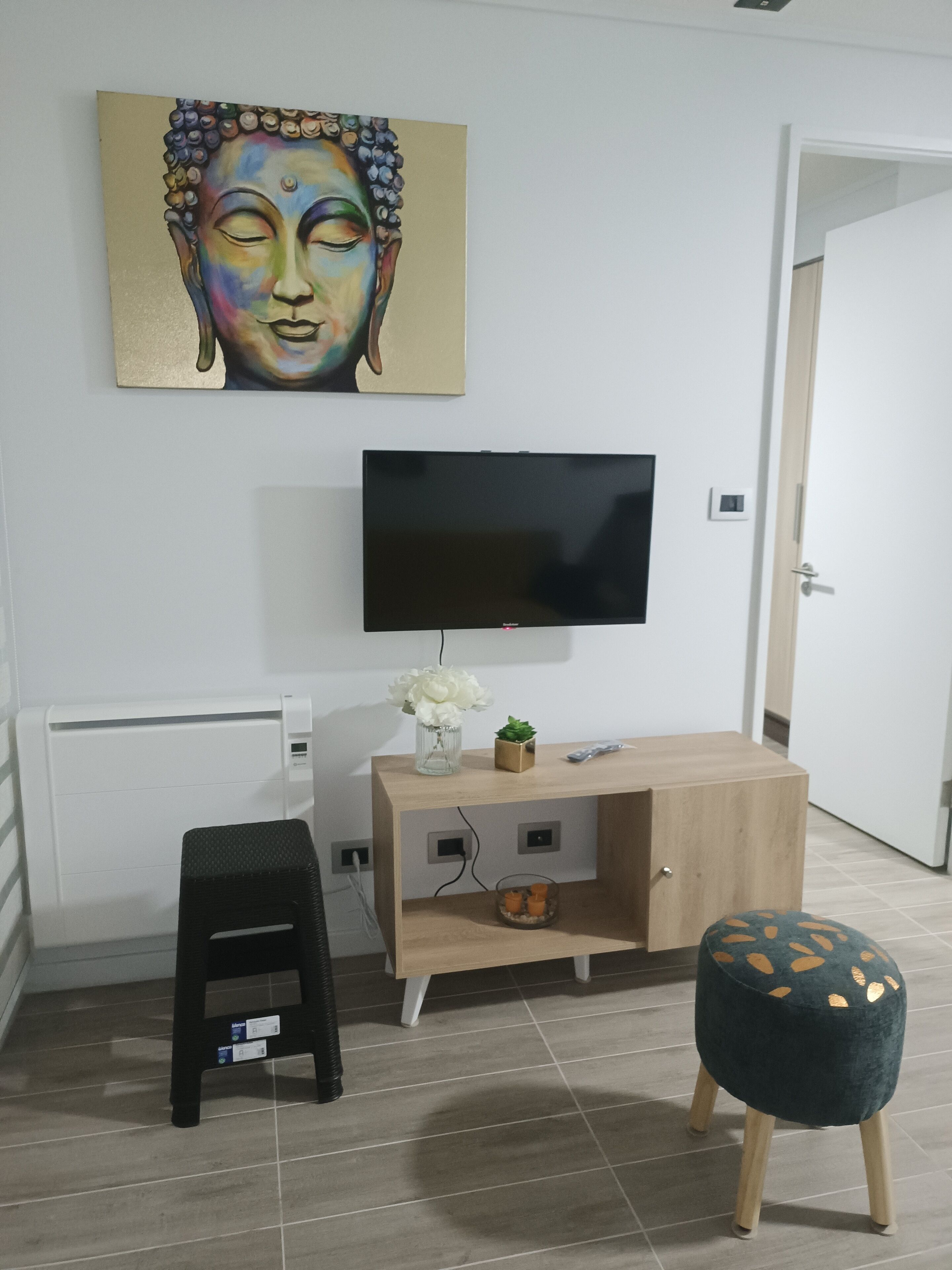 A room with a flat-screen TV mounted on the wall, a small stool, a decorative vase, and a wooden cabinet.