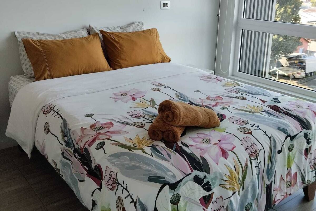 Een slaapkamer met een bed, twee kussens, een raam met zonwering en uitzicht op buiten.